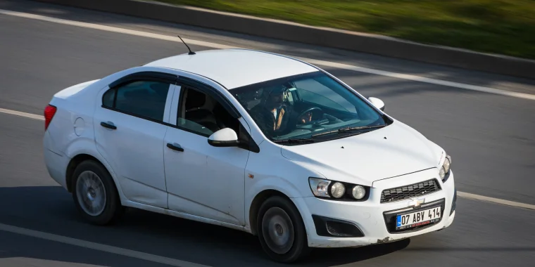 Chevrolet Aveo blanco avanzando rápido en la calle en un día cálido de verano con palmeras al fondo - Seguro de auto barato para el Chevrolet Aveo